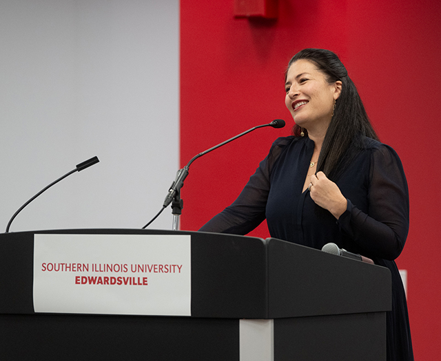 Photography of U.S. Poet Ada Limón reciting a poem at a podium in the legacy room of the Morris University Center. She is wearing a navy dress, has long brown hair, and is smiling as she gestrures toward the audience.